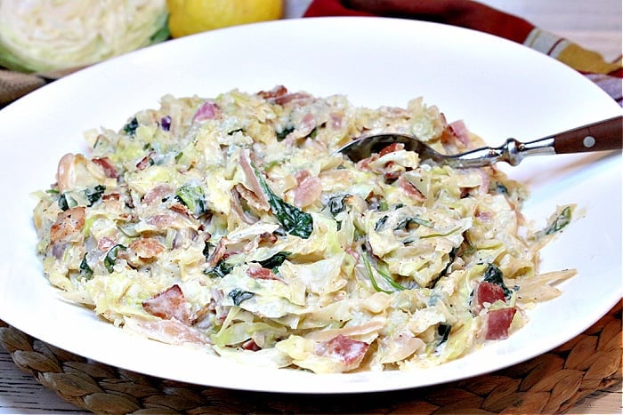 A closeup horizontal photo of a white oblong shaped bowl filled with Creamed Cabbage and Spinach with Bacon and a serving spoon.