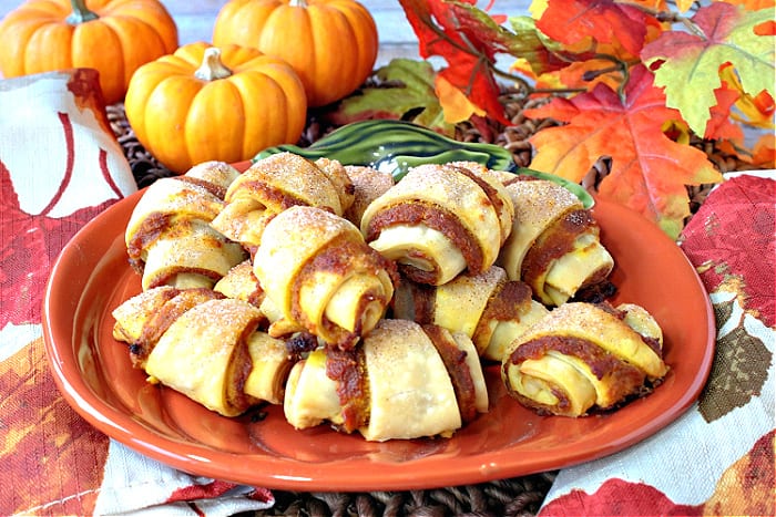 A centered photo of a pumpkin plate filled with Pumpkin Pie Bites with real mini pumpkins in the background along with autumn leaves.