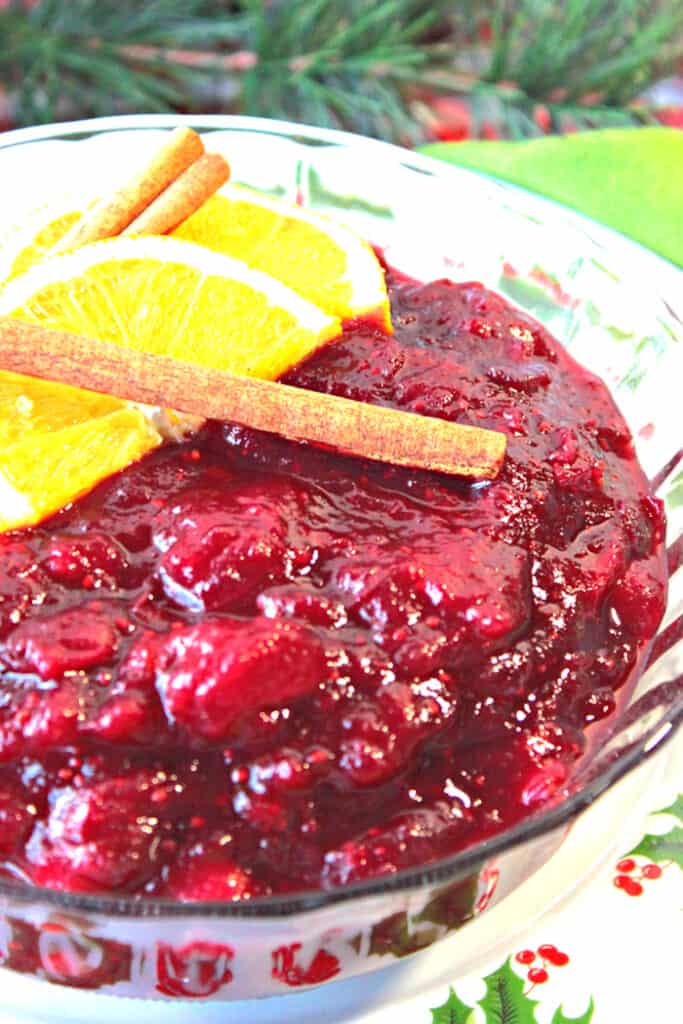 A vertical closeup photo of a glass bowl filled with Whole Berry Cranberry Sauce with cinnamon sticks and orange slices on top