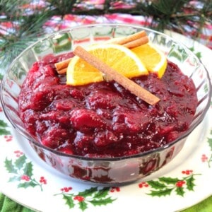 A glass bowl filled with Whole Berry Cranberry Sauce with cinnamon sticks and an orange slice on top.