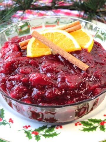 A glass bowl filled with Whole Berry Cranberry Sauce with cinnamon sticks and an orange slice on top.