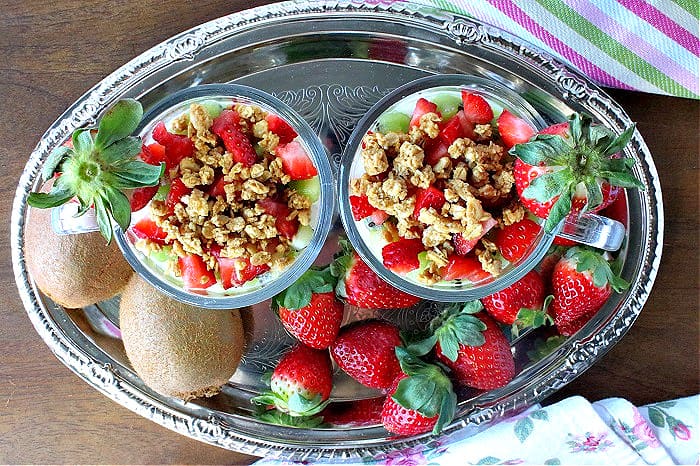 A direct overhead photo of two glass parfait cups filled with Greek yogurt, strawberries, and kiwis on a silver tray.