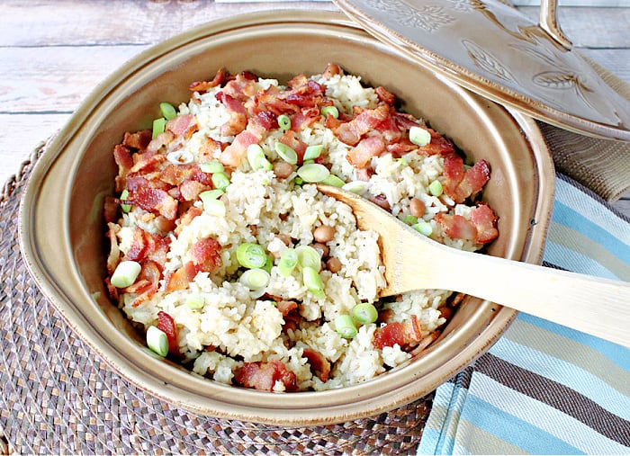 An overhead horizontal photo of Hoppin' John in a round tan casserole dish with scallions and a wooden spoon.