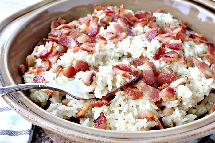 A horizontal closeup of a forkful of Hoppin' John with rice, bacon, and black-eyed peas in a tan casserole dish.