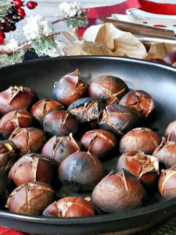 A bunch of cooked Roasted Chestnuts in a chestnut pan along with Christmas colored napkins and an evergreen bough