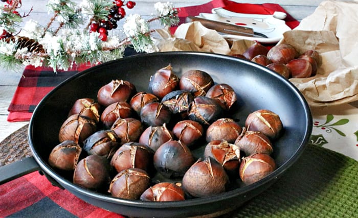 A bunch of cooked Roasted Chestnuts in a chestnut pan along with Christmas colored napkins and an evergreen bough