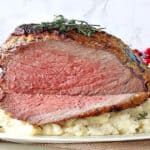 A sliced Whoville Roast Beast on a platter surrounded by mashed potatoes and fresh herbs.