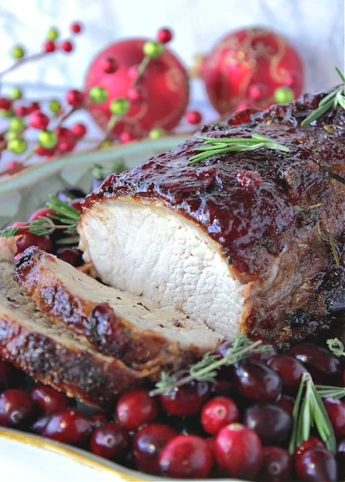 A vertical closeup of a sliced Pork Roast with Cranberry Relish Glaze with fresh herbs and whole cranberries on the platter.