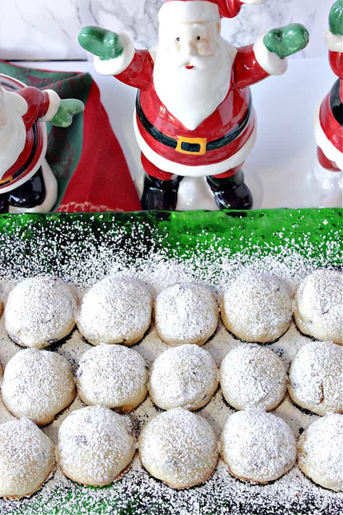 A vertical closeup photo of Snowball Cookies with Pistachios and Cranberries on a green glass plate the some ceramic Santa's in the background.