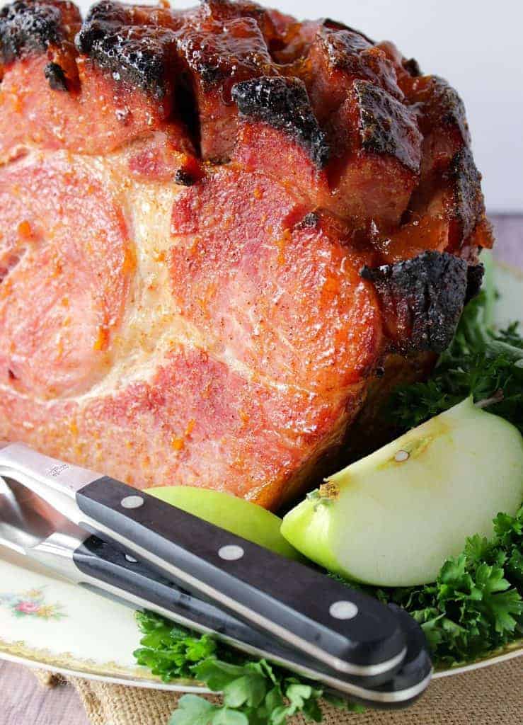 A closeup vertical image of a Spiced Apricot Glazed Ham with a carving set and fresh apple slices.
