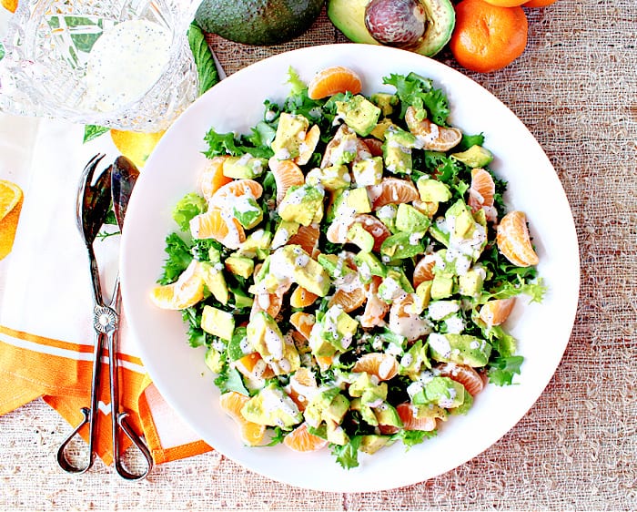 Direct overhead photo of a round white salad bowl filled with retro Avocado Orange Salad drizzled with orange poppy seed dressing.