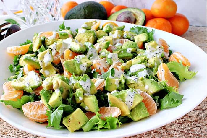 A horizontal photo of a green and orange Avocado Orange Salad in a large white bowl drizzled with poppy seed dressing.