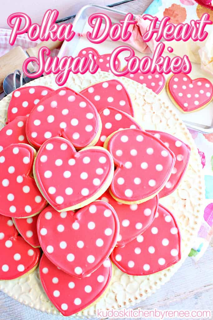 A vertical closeup of a cake plate filled with pink and white Polka Dot Heart Sugar Cookies along with a title text overlay graphic in pink and white.