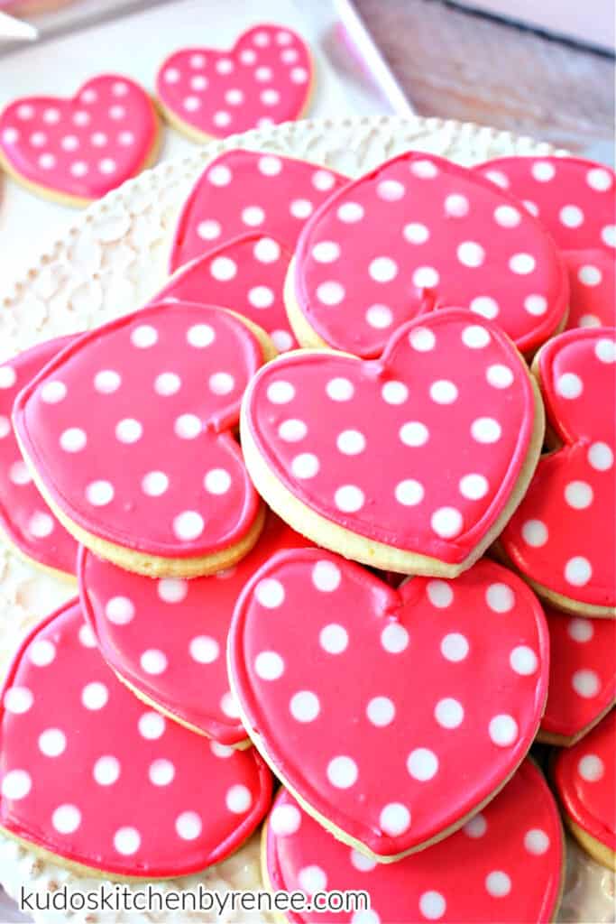 A vertical photo of a stack of Polka Dot Heart Cookies in pink and white along with two cookies on a baking sheet in the background.