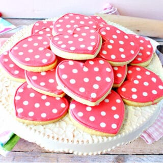 A round cake stand filled with pink Polka Dot Heart Sugar Cookies along with cookie cutters in the background.