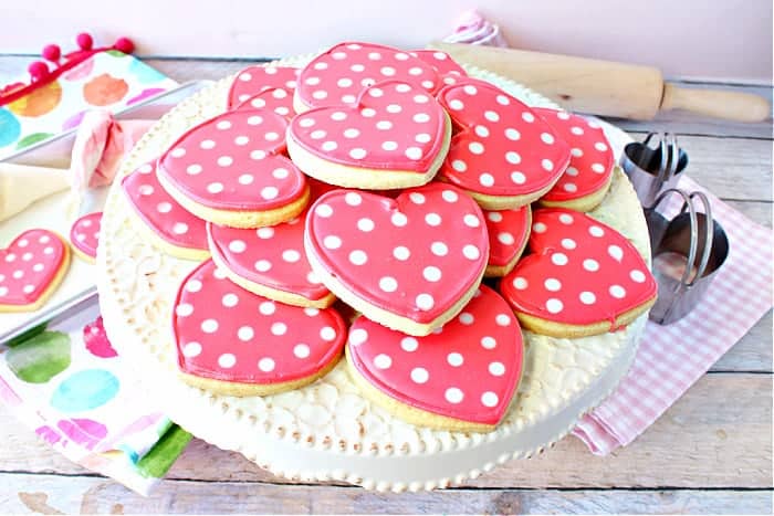 A round cake stand filled with pink Polka Dot Heart Sugar Cookies along with cookie cutters in the background.