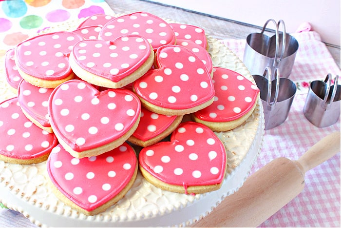 An offset horizontal photo of a cake stand filled with pink Polka Dot Heart Sugar Cookies with a rolling pin in the foreground and cookie cutters in the background.