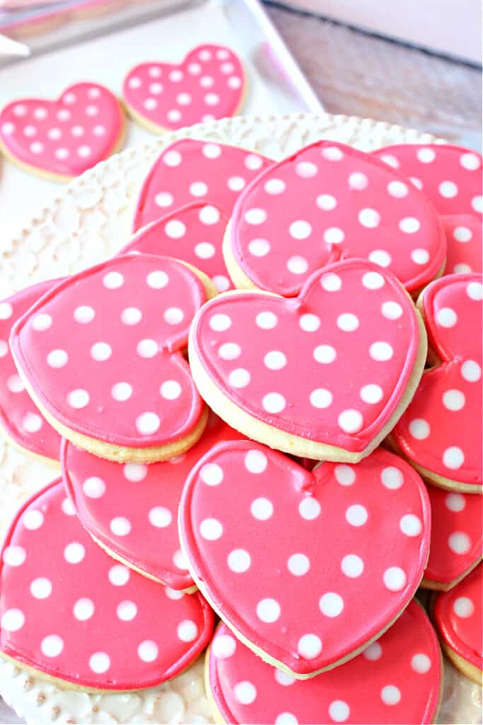 A vertical closeup overhead photo of a pile of pink Polka Dot Sugar Cookies on a platter with a few cookies on a cookie sheet in the background.