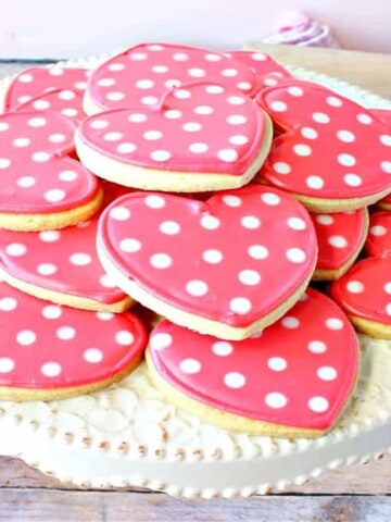 A round cake stand filled with pink Polka Dot Heart Sugar Cookies along with cookie cutters in the background.
