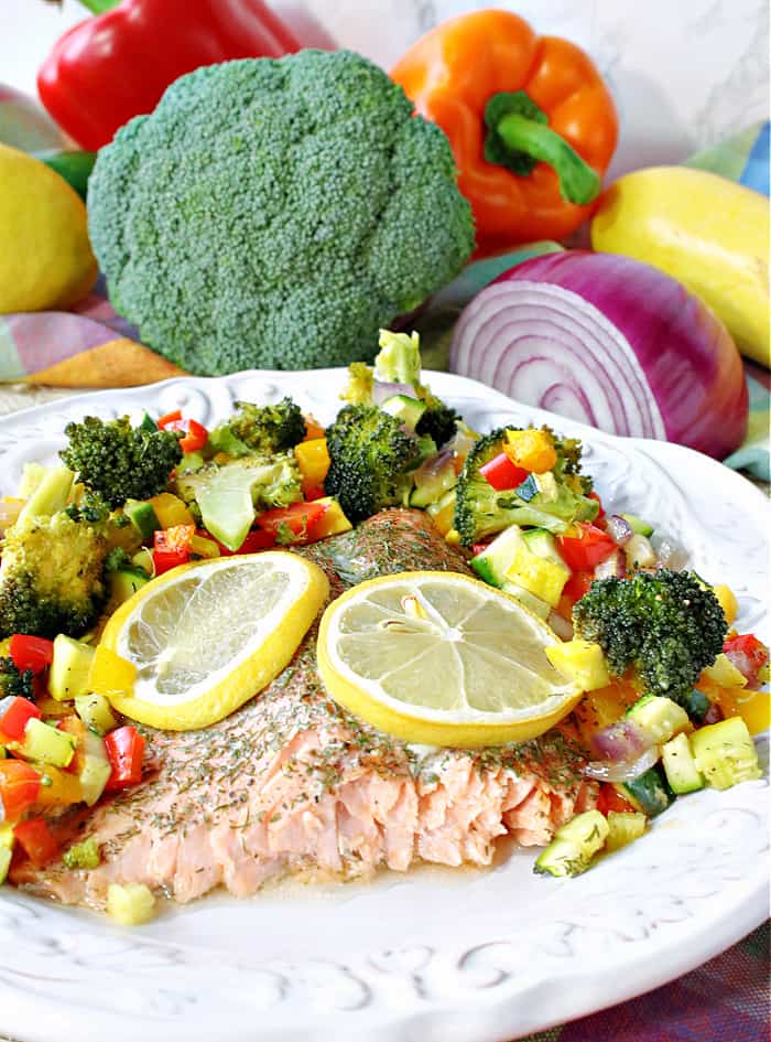 A Rainbow Trout on a white plate surrounded by colorful vegetables and topped with lemon slices.