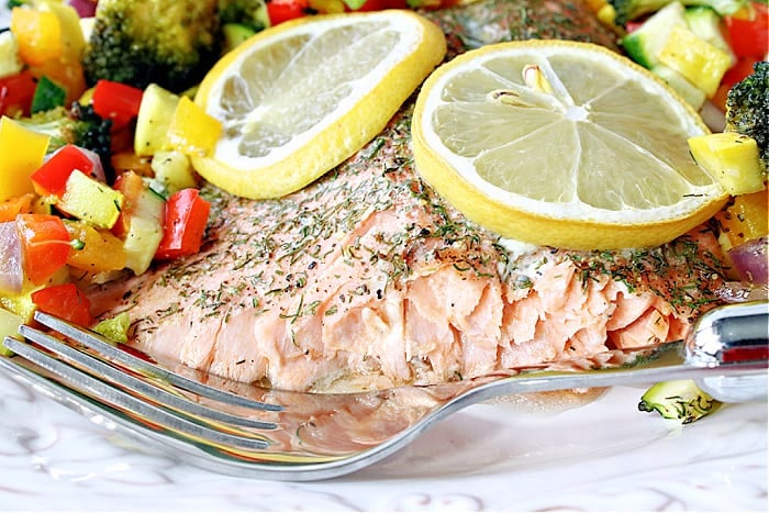 A closeup horizontal photo of the inside of a Roasted Rainbow Trout on a white plate topped with colorful  vegetables and lemon slices.