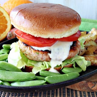 A Miso Salmon Burger on a plate with a bun, tomato, sauce, and sugar snap peas on the side.