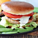 A Miso Salmon Burger on a plate with a bun, tomato, sauce, and sugar snap peas on the side.