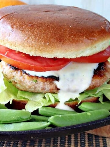 A Miso Salmon Burger on a plate with a bun, tomato, sauce, and sugar snap peas on the side.