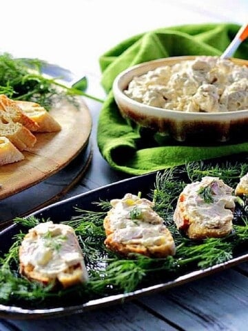 A narrow platter filled with fresh dill sprigs and four Salami and Dill pickle spread crostini.