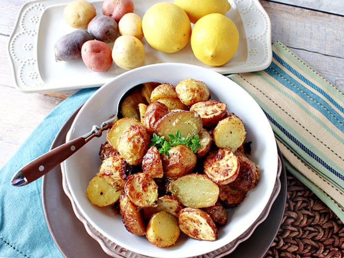 Overhead horizontal photo of Roasted Baby Potatoes with Lemon and Thyme in a bowl with a spoon along with fresh lemons and potatoes in the background.