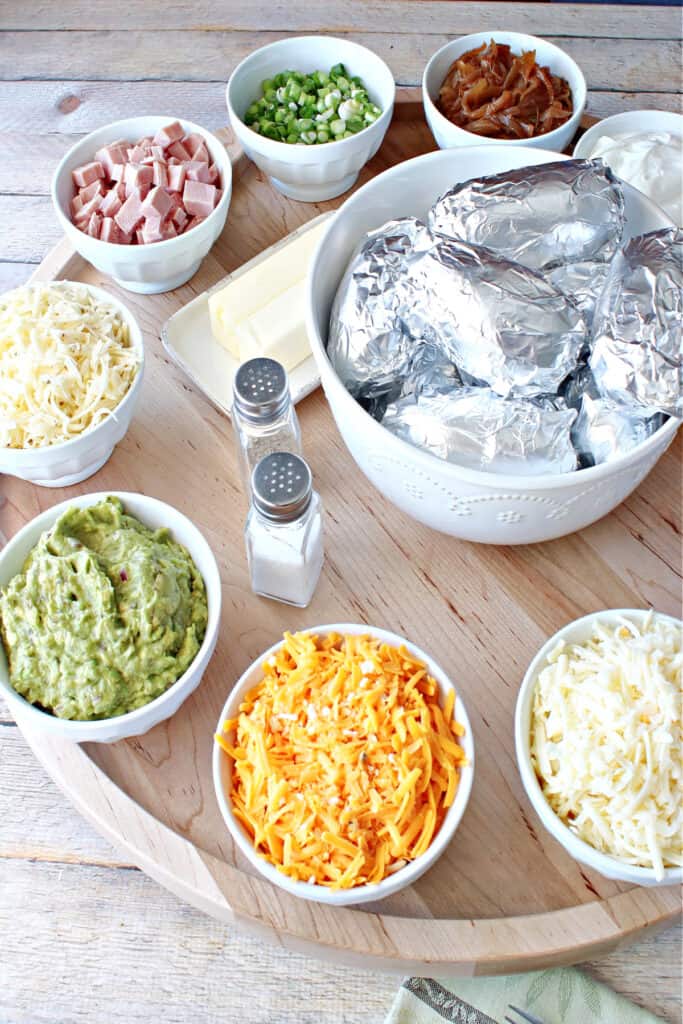 A vertical photo of a Baked Potato Party Board with a bowl of foil wrapped potatoes in the center and potato toppers around the perimeter.