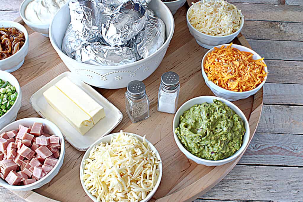 An offset horizontal photo of a large round wooden board with white bowls and topping for serving a Baked Potato Party Board.