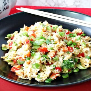 A black bowl of Brown Rice Fried Rice with Vegetables and Eggs sitting on a red plate with chop sticks on the side.