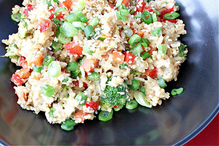 A super closeup horizontal photo of Brown Rice Stir Fry in a black bowl with broccoli, red peppers, scallions and sesame seeds.