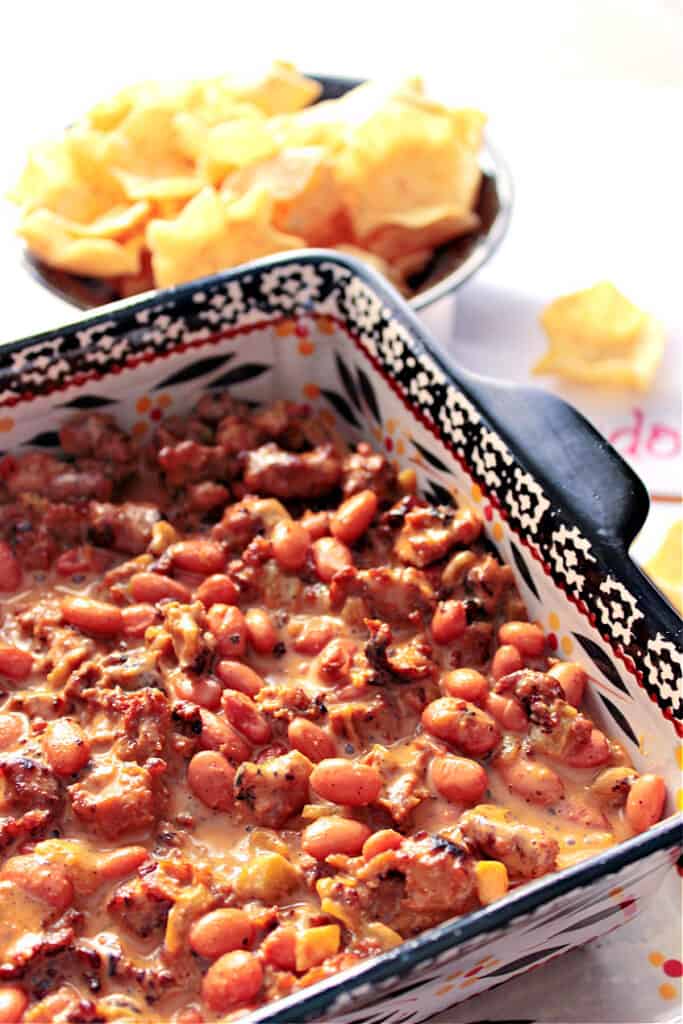 A closeup horizontal photo of a square baking dish filled with Cheesy Sausage Bean Dip and a bowl of chips in the background.