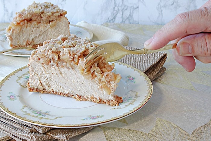 A hand holding a fork and digging into a slice of Dutch Apple Cheesecake on a pretty china plate.