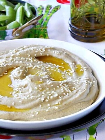 A white bowl filled with Eggplant and Chickpeas Hummus in the foreground and a small bowl of sugar snap peas in the background.
