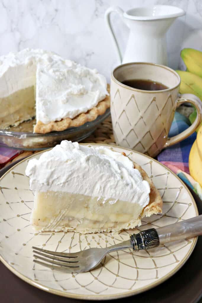 A slice of Mom's Best Banana Cream Pie on a plate with a fork with a cup of coffee and the rest of the pie in the background.