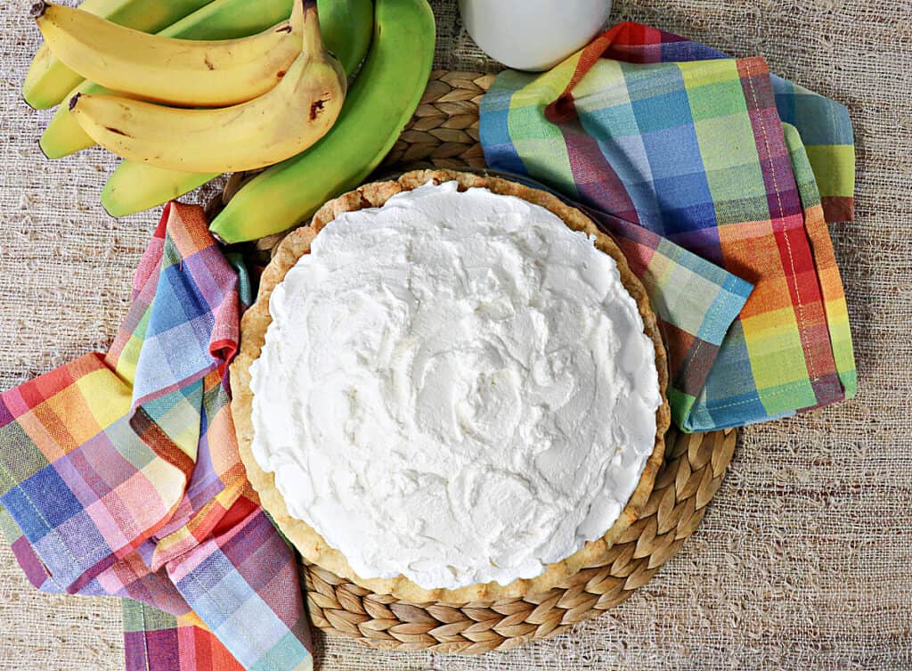 An overhead photo of an entire banana cream pie topped with whipped cream with fresh bananas in the background.