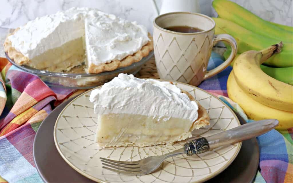 A slice of Mom's Best Banana Cream Pie on a plate with a fork along with a cup of coffee and the rest of the pie in the background.