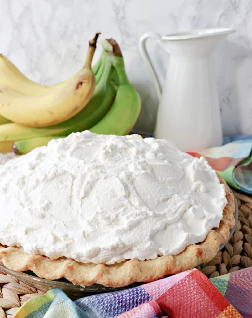 A vertical photo of Mom's Best Banana Cream Pie loaded with whipped cream with fresh bananas in the background.