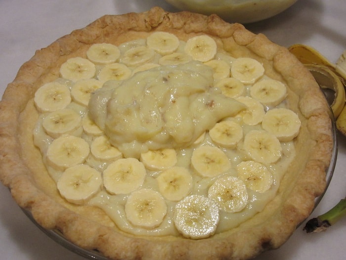 Banana custard being poured over a second layer of sliced bananas for a pie.