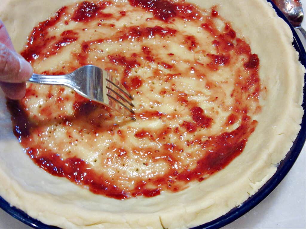 A closeup horizontal photo of Mom's Homemade Pie Crust in a blue pie plate with a layer of strawberry jam and a fork.
