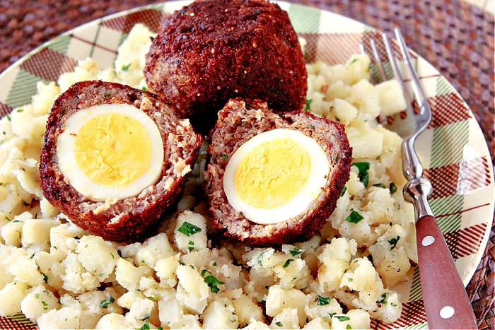A closeup horizontal photo of two Scotch Eggs on a plaid plate with potatoes and a fork.