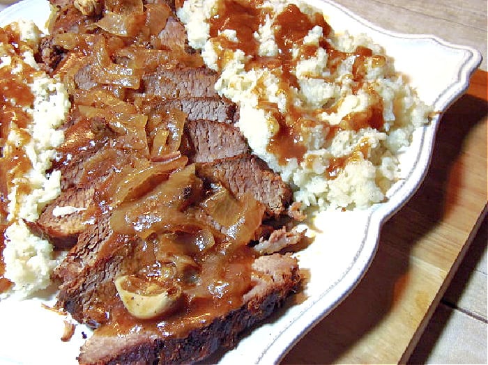 A closeup photo of a sliced Beef Brisket on a square white plate with mashed potatoes, gravy, and onions.