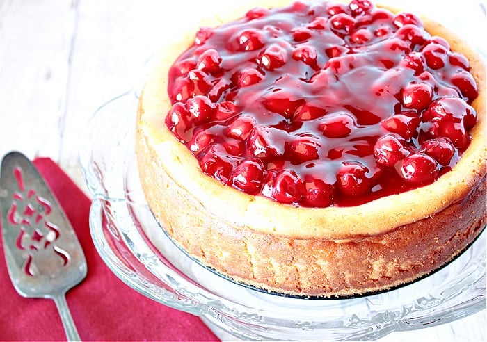 An offset horizontal photo of a cherry cheesecake on a glass plate with a pie server and a red napkin in the background.