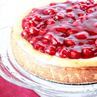 An offset horizontal photo of a Traditional Cherry Cheesecake on a glass cake stand with a cake server and a red napkin.