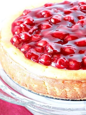An offset horizontal photo of a Traditional Cherry Cheesecake on a glass cake stand with a cake server and a red napkin.