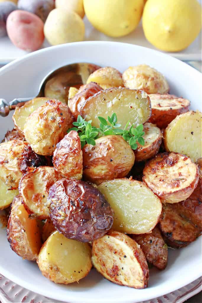 A closeup vertical image of golden brown Air Fryer Baby Potatoes in a white bowl with lemons and baby potatoes in the background.