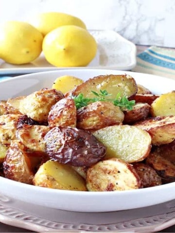 A horizontal photo of a white bowl filled with Air Fryer Baby Potatoes with fresh lemons and potatoes in the background.